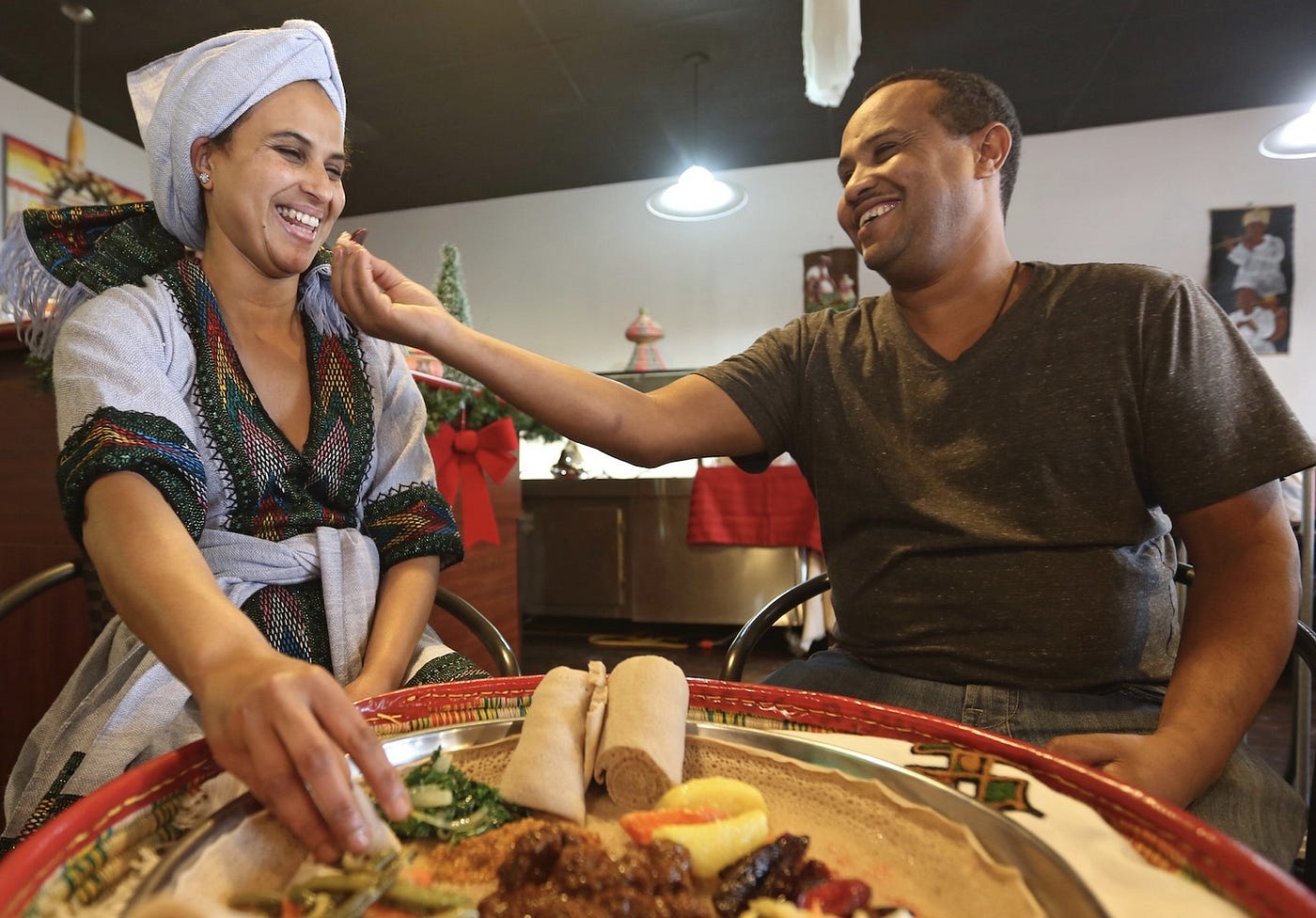 A man reaches across a shared plate of injera to place a gursha — a bite of food — into a smiling woman's mouth with his hand.